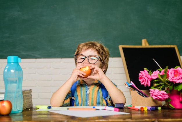 Cute boy at lunch time in classroom young schoolboy sitting at table with apple in class room little