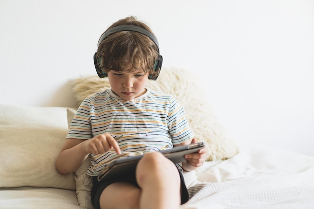 Ragazzo carino sdraiato sul letto a casa e giocare con il tablet pc o leggere online e ascoltare musica con le cuffie wireless