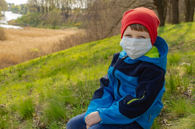 Cute boy in a medical mask sits on a hill on a log and look at the lake. Family walks with children outdoors in early spring during quarantine