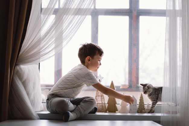 Cute boy playing with kitten sitting on the windowsill near the window. Cozy home