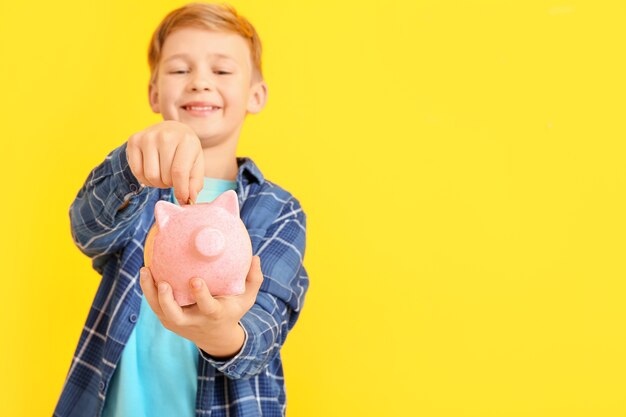 Cute boy putting money in piggy bank on color background