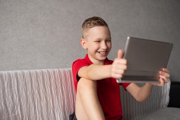 A cute boy in a red Tshirt is sitting playing on a tablet and laughing