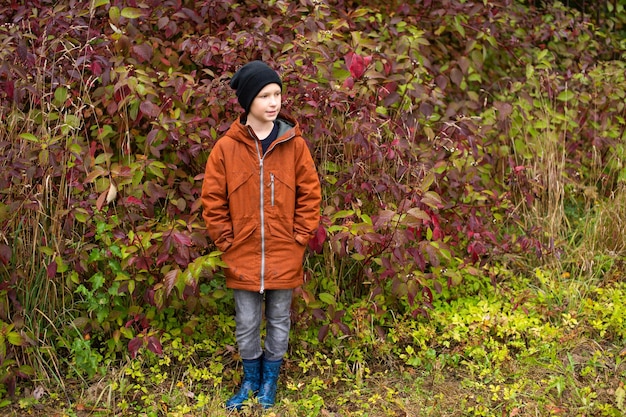 A cute boy in rubber boots stands in the autumn forest