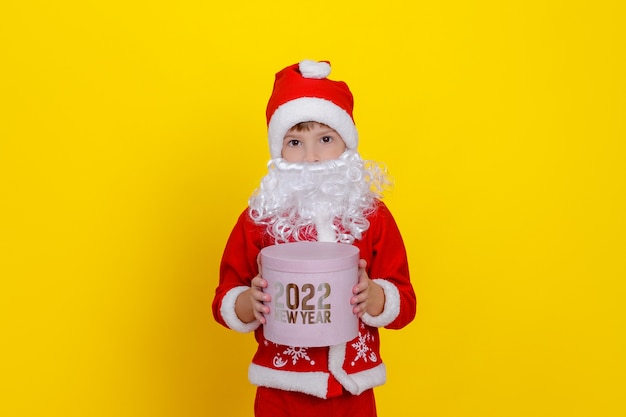 A cute boy in Santa Claus clothes and with an artificial white beard holds a pink round gift box