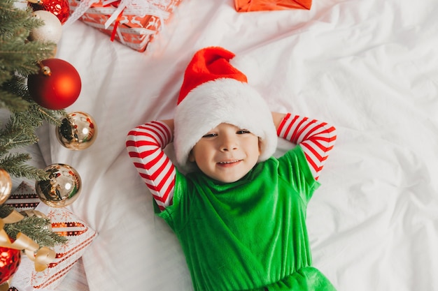 Cute boy in santa claus hat lies on a blanket near the christmas tree. happy child lies near the Christmas tree and smiles