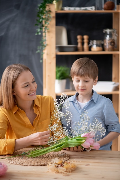 Ragazzo carino in camicia in piedi al tavolo e aiutare la madre a organizzare bouquet di tulipani e respiro del bambino in cucina