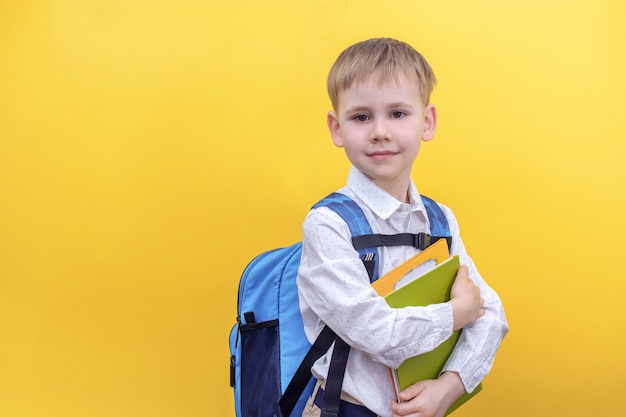 A cute boy in a shirt with a backpack on his back holding textbooks on yellow