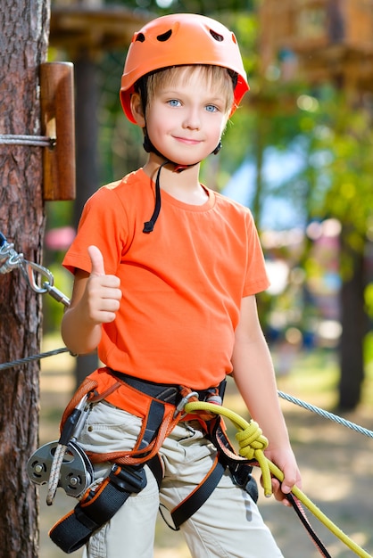 かわいい男の子がアドベンチャーパークの登山用具で親指を表示します。