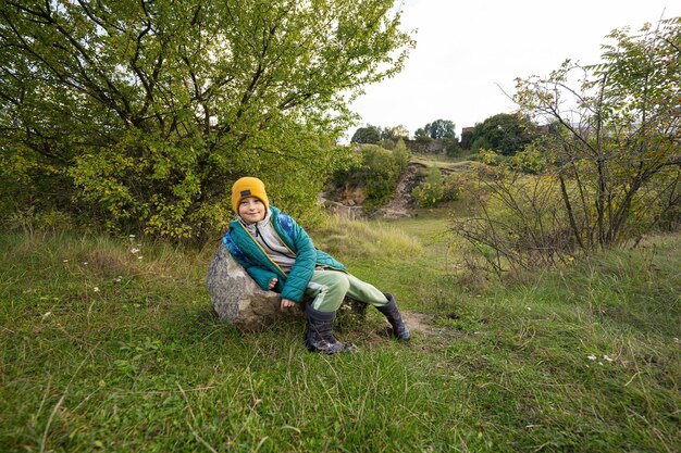 Cute boy sit on stone at autumn hill wearing a green jacket yellow hat with backpack and rubber boots