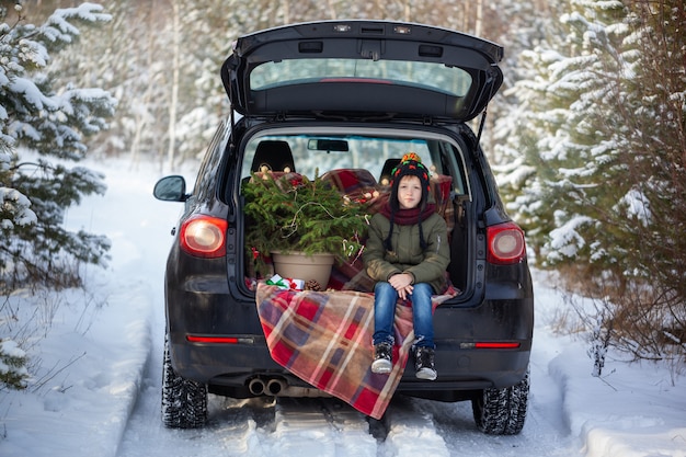 Cute boy sitting in black car at snowly winter forest. Christmas concept.