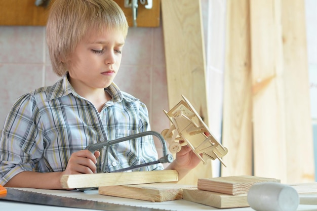 Cute boy sitting at table with model of ship and handsaw and working with wooden planks