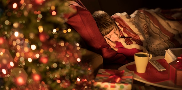 Милый мальчик спит в гостиной рядом с елкой и ждет Санту в канун Рождества.