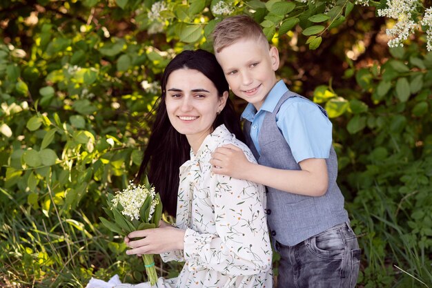 A cute boy stands and hugs his mother who has a bouquet of lilies of the valley in her hands