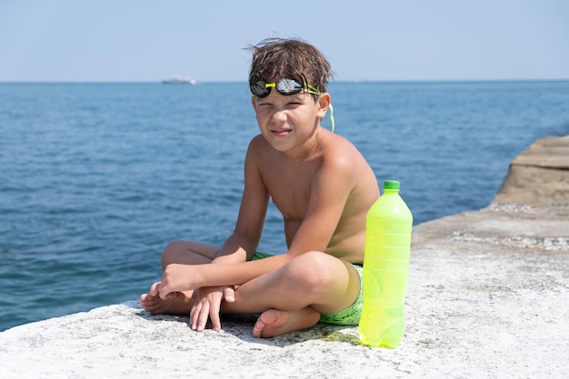 A cute boy in swimming glasses and shorts with a bottle of water sits on a pier on the seashore.