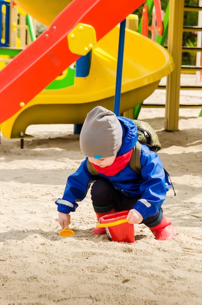 Милый двухлетний мальчик в синем пиджаке и красном шарфе играет на яркой игровой площадке с ковшом и ведром
