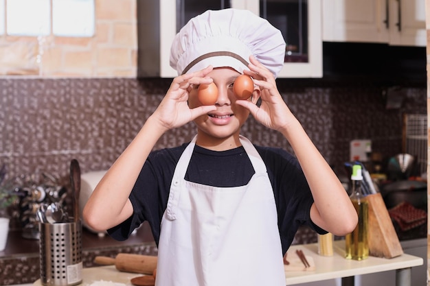 Милый мальчик в униформе шеф-повара кладет яйца на глаза, взволнованный готовкой на кухне