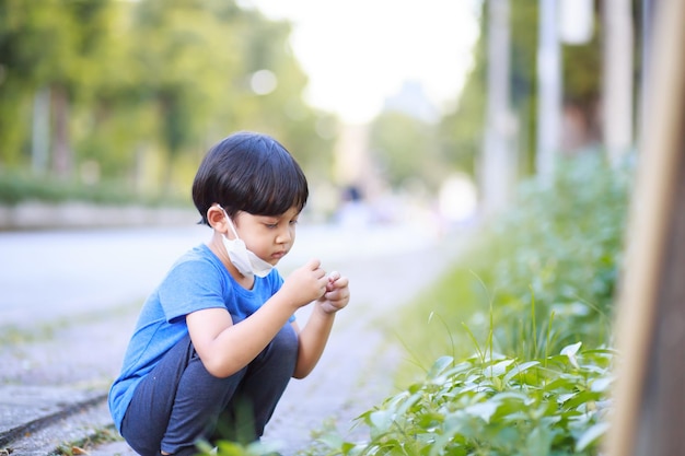 Симпатичный мальчик в синей рубашке, длинных черных штанах и белой маске сидит весной в пышном зеленом саду на фоне зеленых деревьев.