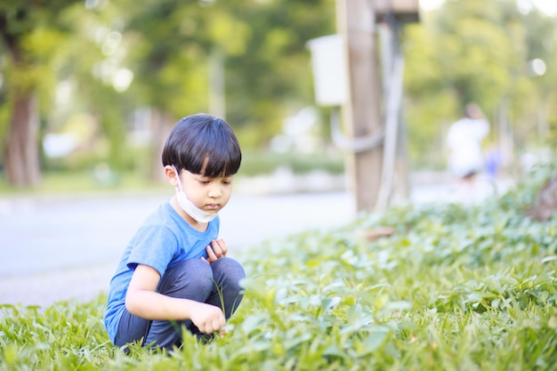 青いシャツの長い黒のズボンと白いマスクを身に着けているかわいい男の子は、緑の木々のポートレートショットを背景に春に緑豊かな庭園に座っています