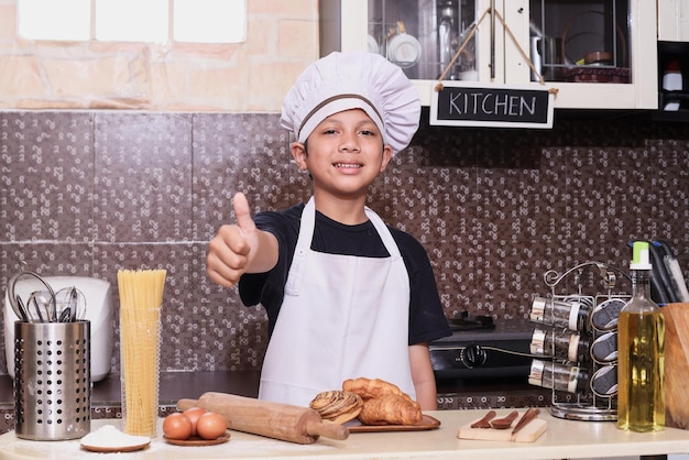 シェフのエプロンを着たかわいい男の子が、キッチンで焼きたてのパンを見せながら親指を上げます。