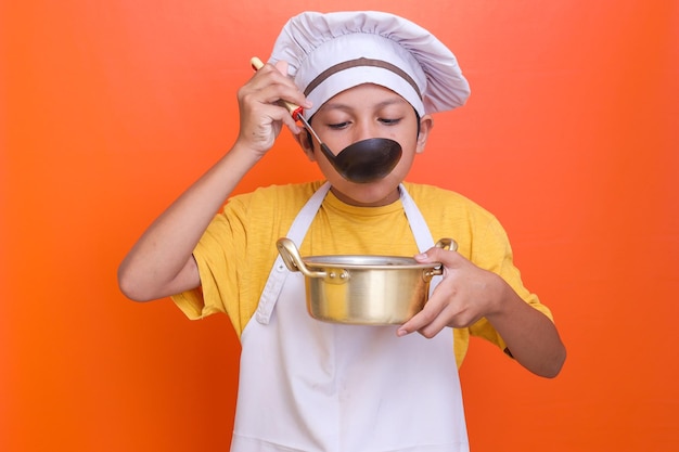 Cute boy wearing chef uniform and apron tasting soup with black ladle isolated orange backgroundd