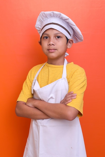 요리사 유니폼을 입은 귀여운 소년은 주황색 배경에 자신 있게 고립되어 있습니다.