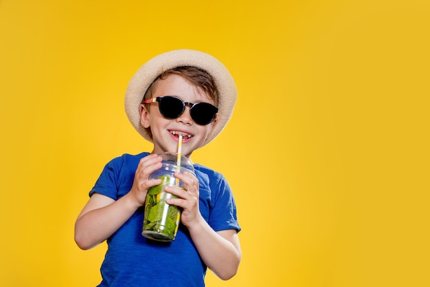 かわいい男の子はサングラスをかけ、プラスチック製のコップでレモネードを飲みます