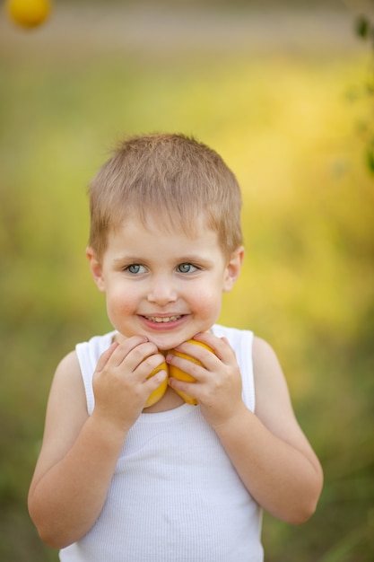 A cute boy with blond hair in a white T-shirt with summer lemons in the garden under a tree