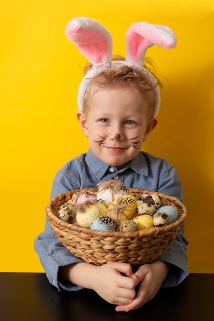 Cute boy with bunny ears holding basket with easter eggs on yellow background copy space