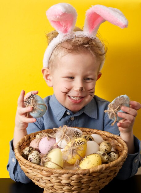 Cute boy with bunny ears holding basket with easter eggs on yellow background copy space