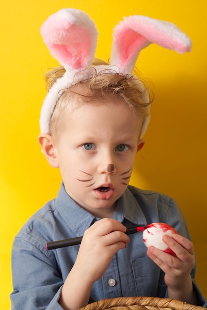 Cute boy with bunny ears paints easter eggs on a yellow background