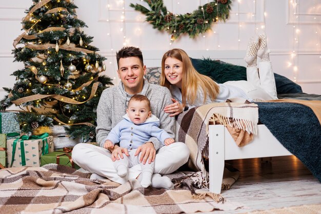 Cute boy with happy parents near the Christmas tree A family with a small child in a bedroom near a decorated Christmas tree Festive New Year's atmosphere Family celebration