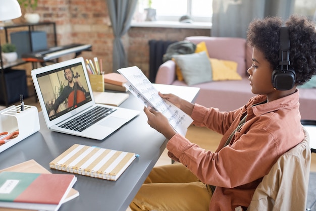 Photo cute boy with papers with musical notes sitting in front of laptop in home environment during online lesson