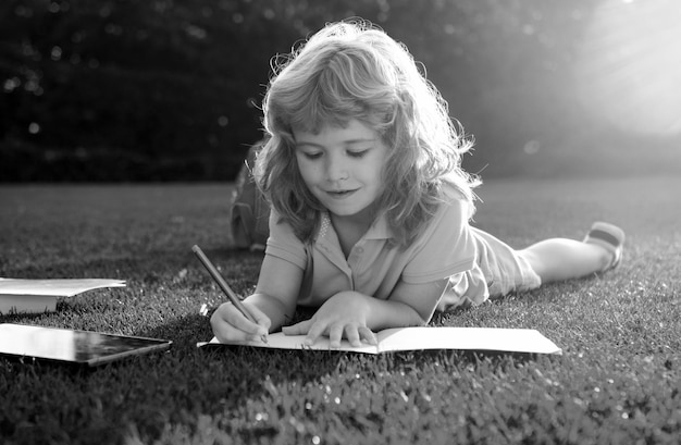 Cute boy with pencil writing on notebook laying on grass Child reading a book in the summer park