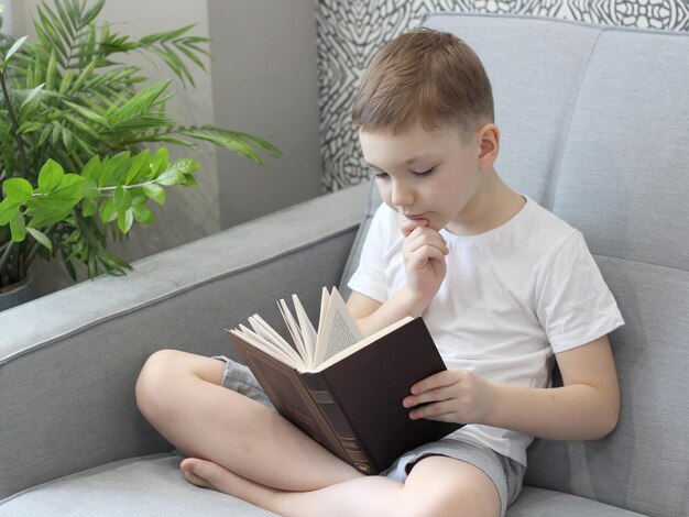 Cute boyin a white tshirt is sitting on the sofa and reading a book the book is upside down