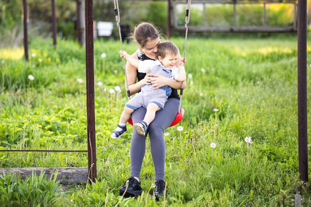 Cute brother and older sister ride on a bright swing on warm spring day against the backdrop of a green blooming garden