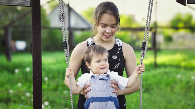 Cute brother and older sister ride on a bright swing on warm spring day against the backdrop of a green blooming garden