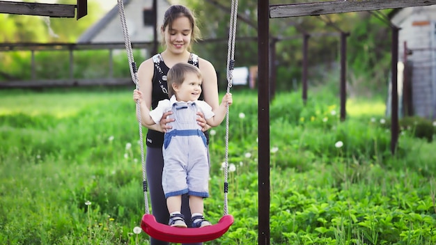 Cute brother and older sister ride on a bright swing on warm spring day against the backdrop of a green blooming garden