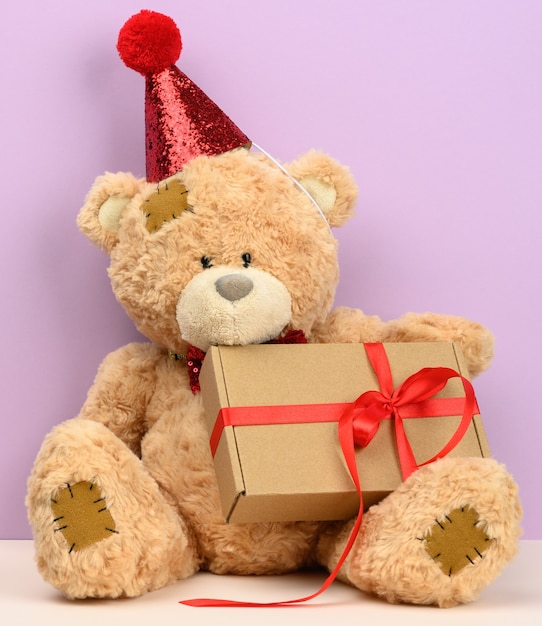Cute brown teddy bear in a red cap sits and holds a brown box with a gift, festive surface