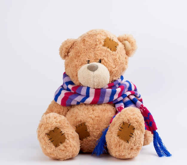 Cute brown teddy bear with patches in a colored knitted scarf sitting