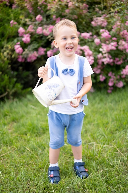 Cute brownhaired baby with blue eyes in summer a bright sunny day the child laughs holds a watering can waters flowers taking care of nature