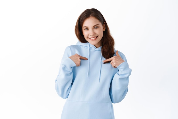 Cute brunette girl in casual hoodie pointing at herself, smiling and self-promoting, choose me gesture, standing against white background