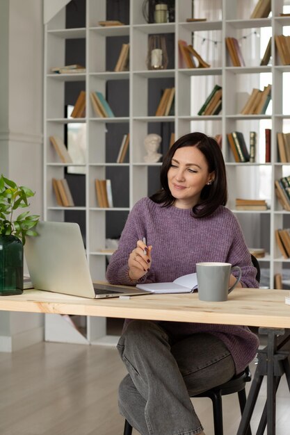 a cute brunette girl in a lilac sweater is sitting at a table with a laptop making notes