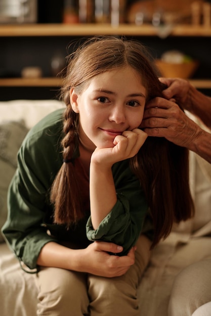 Cute brunette girl looking at camera while her grandmother plaiting pigtail