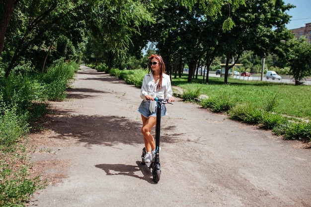 Cute brunette girl rides on an electrode scooter in the park she is wearing a white shirt. concept of eco transport and rent.