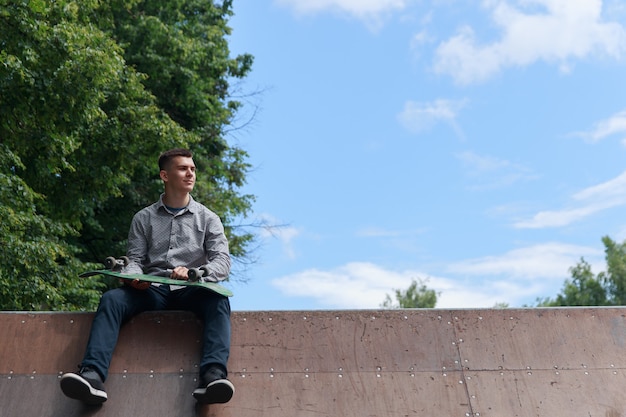 Cute brunette guy looking to the side and holding skateboard sitting on skateboard slide in the park on blue sky background on hot summer day, copyspace