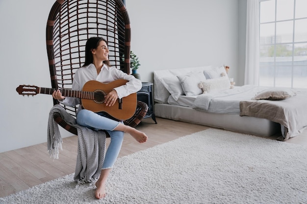 Foto bruna carina in camicia bianca e jeans blu seduto su una sedia sospesa di vimini a suonare la chitarra