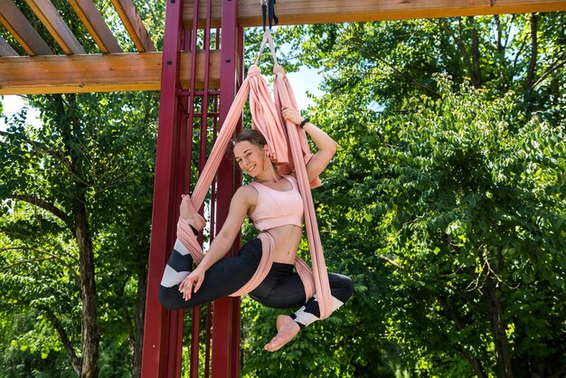 Cute brunette with gorgeous hair in dark leggings stretches beautifully on the canvases that are attached to the tree The concept of stretching outdoors in good weather in good weather