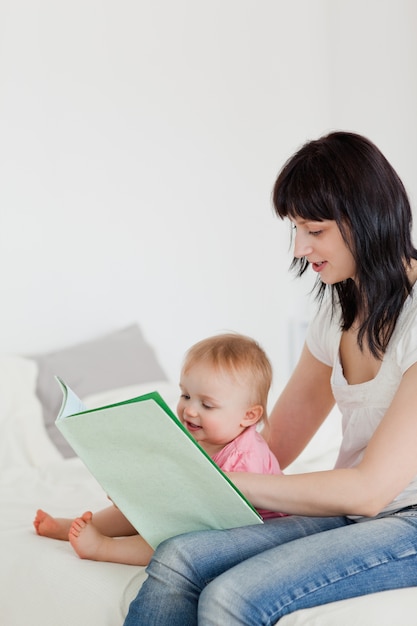 Cute brunette woman showing a book to her baby while sitting on a bed