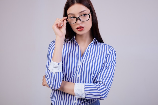 Cute business woman with brown hair, full pink lips posing. She is wearing a white and blue striped shirt and black computer glasses. Worker.