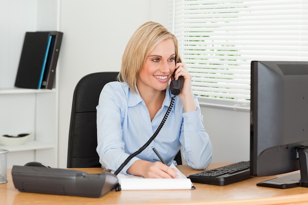 Cute businesswoman on phone writing something down looks at screen